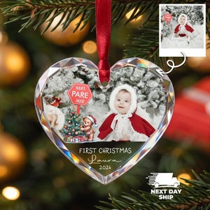 K&ouml;nnte beinhalten: Ein herzf&ouml;rmiger Kristallanh&auml;nger mit rotem Band, der ein Baby mit rotem Umhang und wei&szlig;er Kapuze, einen Teddyb&auml;ren, einen Weihnachtsbaum und ein rotes Schild mit der Aufschrift NOEL PARE AQUI zeigt. Text: FIRST CHRISTMAS Laura 2026.