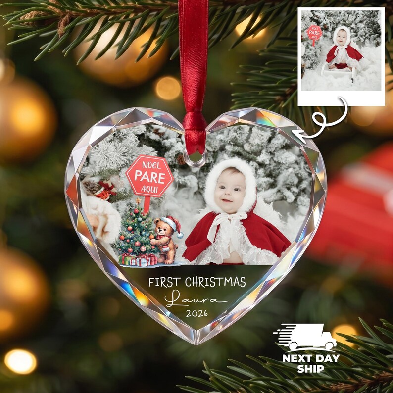 K&ouml;nnte beinhalten: Ein herzf&ouml;rmiger Kristallanh&auml;nger mit rotem Band, der ein Baby mit rotem Umhang und wei&szlig;er Kapuze, einen Teddyb&auml;ren, einen Weihnachtsbaum und ein rotes Schild mit der Aufschrift NOEL PARE AQUI zeigt. Text: FIRST CHRISTMAS Laura 2026.