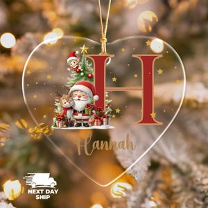May include: Heart-shaped Christmas ornament with a festive design. The ornament features a red letter "H" and a Santa Claus illustration with a reindeer, gifts, and a Christmas tree. The name "Hannah" is written in gold script.