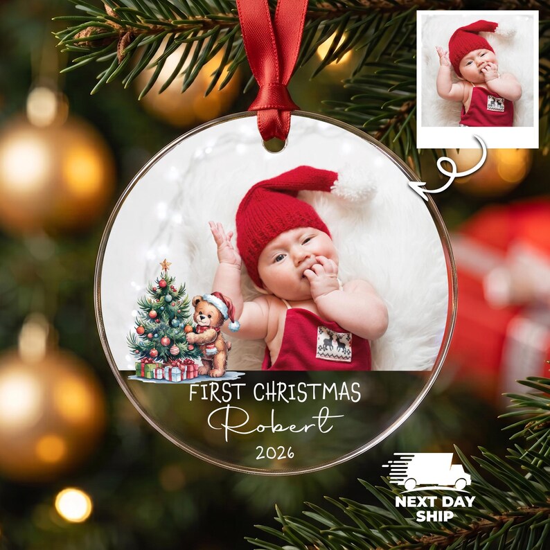 K&ouml;nnte beinhalten: Ein runder, transparenter Acryl-Weihnachtsbaumschmuck mit einem Foto eines Babys in roter M&uuml;tze und roter Latzhose, einem kleinen Teddyb&auml;ren neben einem geschm&uuml;ckten Weihnachtsbaum und dem Text First Christmas Robert 2026 mit rotem Band.