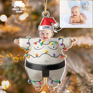 May include: A personalised Christmas ornament featuring a baby's face wearing a Santa hat with the name "Liam". The ornament is shaped like a sumo wrestler wearing a white inflatable suit decorated with colourful Christmas lights.