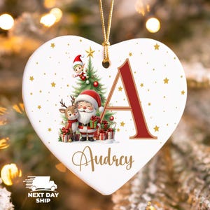 May include: Heart-shaped Christmas ornament with a festive design. Features Santa Claus, a reindeer, and an elf near a Christmas tree. The letter "A" is in red, with the name "Audrey" below. Gold stars and a gold hanging cord complete the design.