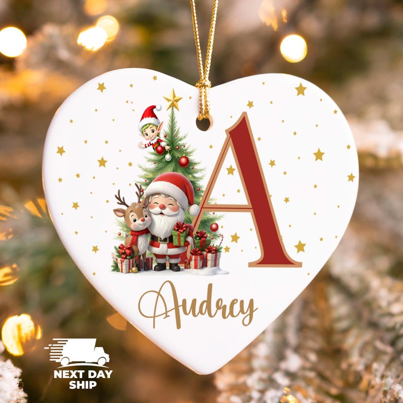 May include: Heart-shaped Christmas ornament with a festive design. Features Santa Claus, a reindeer, and an elf near a Christmas tree. The letter "A" is in red, with the name "Audrey" below. Gold stars and a gold hanging cord complete the design.