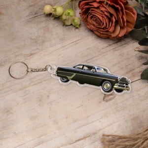 Pu&ograve; includere: Un portachiavi di auto vintage nero con un anello portachiavi argentato. L'auto &egrave; un modello degli anni '50 con un paraurti cromato e pneumatici a fascia bianca.