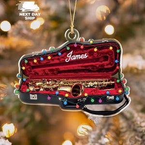 May include: A Christmas ornament shaped like a saxophone in its case, with the name "James" in white script. The case is black with a red velvet interior, and the ornament is decorated with colorful string lights.
