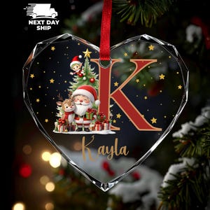 May include: Heart-shaped Christmas ornament with a red ribbon. The ornament features a festive design with Santa Claus, a reindeer, and a Christmas tree. A large red letter "K" and the name "Kayla" are also displayed.