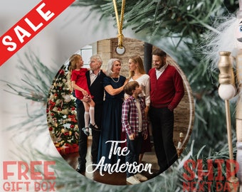 May include: A round ceramic ornament featuring a printed photograph of a family standing together in a room with a Christmas tree. The text The Anderson is printed in white script at the bottom center. A gold cord is attached to the top hole.