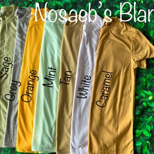 Op de afbeelding: Zes verschillende gekleurde T-shirts die op een groene, bladerige achtergrond liggen. De kleuren zijn grijsgroen, oranje, mint, bruin, wit en karamel. De tekst "Nosagb's Blanks" staat bovenaan de afbeelding.