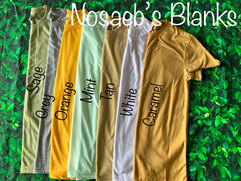 Op de afbeelding: Zes verschillende gekleurde T-shirts die op een groene, bladerige achtergrond liggen. De kleuren zijn grijsgroen, oranje, mint, bruin, wit en karamel. De tekst "Nosagb's Blanks" staat bovenaan de afbeelding.