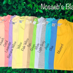 May include: A row of 14 blank t-shirts in various colors, including pink, peach, coral, lavender, yellow, pale yellow, tan, green, white, light grey, aqua, blue, and mustard. The text "Nosaeb's Blanks" is at the top of the image.