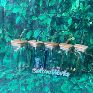 May include: Five clear glass tumblers with bamboo lids and matching colored straws. The straws are blue, green, yellow, and orange. The tumblers have a handle on one side. The tumblers are arranged in a row on a green leafy background. The text "@mosaebblanks" is visible in the bottom center of the image.