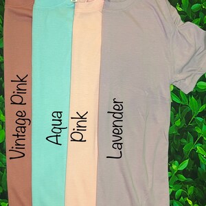 Op de afbeelding: Vier effen T-shirts in verschillende kleuren: vintage roze, aqua, roze en lavendel. De shirts liggen naast elkaar op een groene, bladerrijke achtergrond. Elk shirt heeft een klein label met de merknaam "Nosaleb's" op de voorkant.