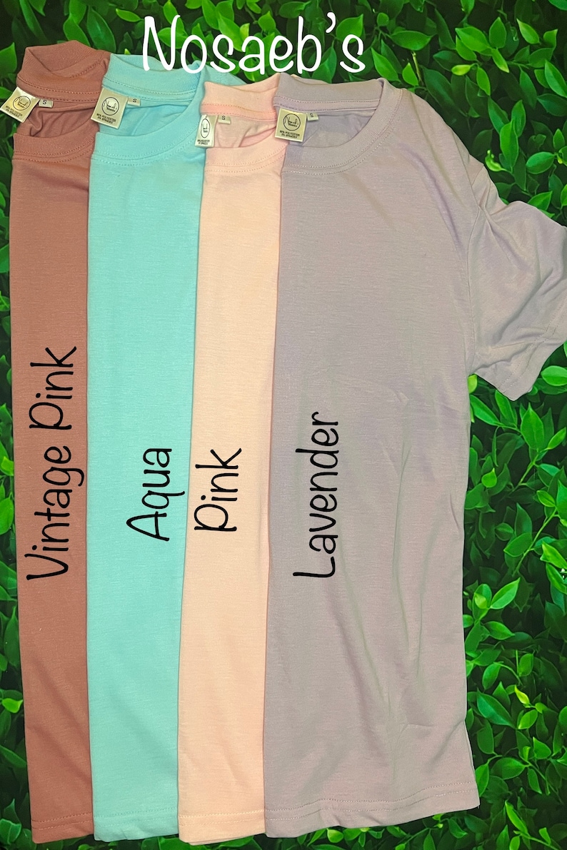 Op de afbeelding: Vier effen T-shirts in verschillende kleuren: vintage roze, aqua, roze en lavendel. De shirts liggen naast elkaar op een groene, bladerrijke achtergrond. Elk shirt heeft een klein label met de merknaam "Nosaleb's" op de voorkant.
