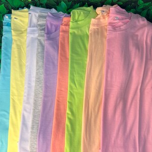 Puede incluir: Una pila de diez camisetas lisas en varios colores pastel, incluyendo azul claro, amarillo, gris, lavanda, melocot&oacute;n, verde y rosa.
