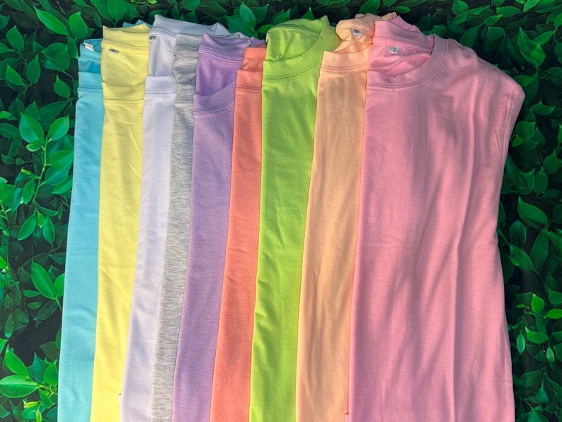 Puede incluir: Una pila de diez camisetas lisas en varios colores pastel, incluyendo azul claro, amarillo, gris, lavanda, melocot&oacute;n, verde y rosa.