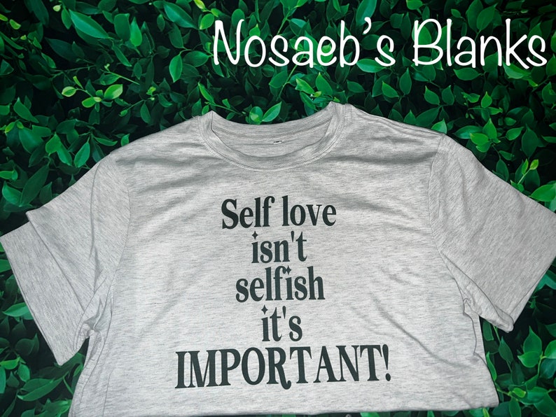 Puede incluir: Una camiseta gris con el texto "Self love isn't selfish it's IMPORTANT!" impreso en negro.