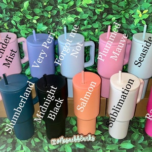 Op de afbeelding: Een verzameling van 12 kleurrijke roestvrijstalen bekers met handvatten en rietjes. De bekers zijn in twee rijen gerangschikt, waarbij de bovenste rij kleuren zoals lavendel, blauw en roze toont en de onderste rij kleuren zoals zwart, oranje en wit. De bekers zijn gelabeld met namen zoals "Winter White", "Lavender Mist", "Very Peri", "Forgot me Not", "Plummed Mauve", "Seaside", "Redwood", "Twirled in Tulle", "Slumberland", "Midnight Black", "Salmon", "Sublimation", "Raspberry Vibes".
