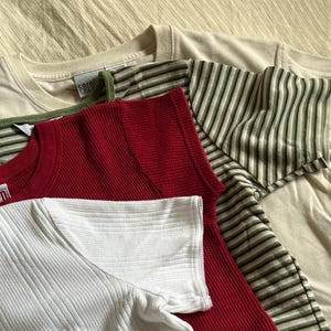 May include: An assortment of folded t-shirts in various colors and textures. The shirts include a cream-colored tee, a red waffle-knit long-sleeve, a white ribbed short-sleeve, and a striped green and cream tee. The "carol cole" brand tag is visible on one shirt.