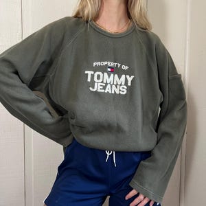 May include: Olive green sweatshirt with the text "PROPERTY OF TOMMY JEANS" in white. The sweatshirt is paired with navy blue athletic shorts with a white drawstring and white stripes on the sides. The outfit is casual and sporty.