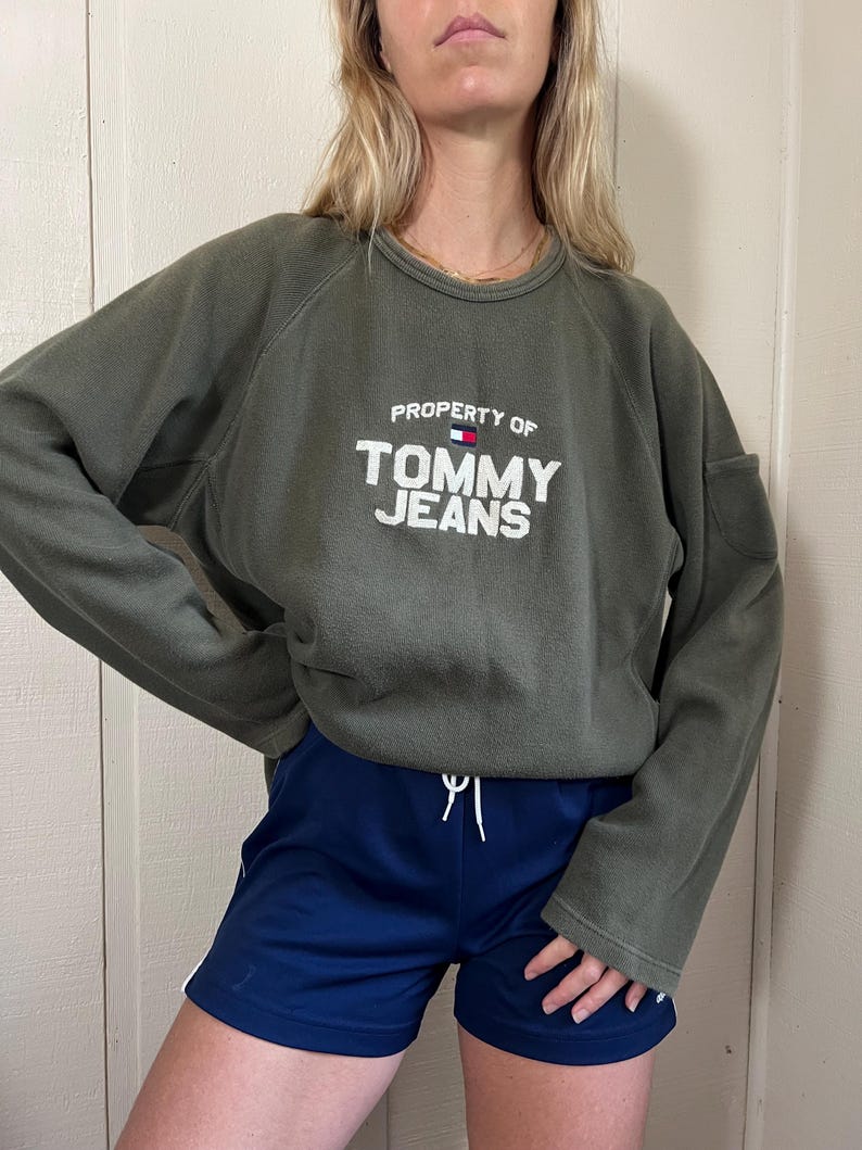 May include: Olive green sweatshirt with the text "PROPERTY OF TOMMY JEANS" in white. The sweatshirt is paired with navy blue athletic shorts with a white drawstring and white stripes on the sides. The outfit is casual and sporty.