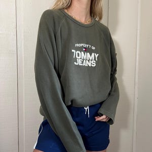May include: Olive green sweatshirt with the text "PROPERTY OF TOMMY JEANS" in white. The sweatshirt is paired with navy blue shorts with a white stripe down the side. The outfit is casual and sporty.