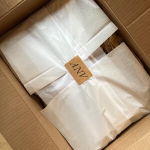 K&ouml;nnte beinhalten: Ein offener Karton mit verpackten Artikeln. Die Artikel sind in wei&szlig;es Seidenpapier eingewickelt und mit einem kleinen braunen Etikett mit der Aufschrift "ANV" versehen.
