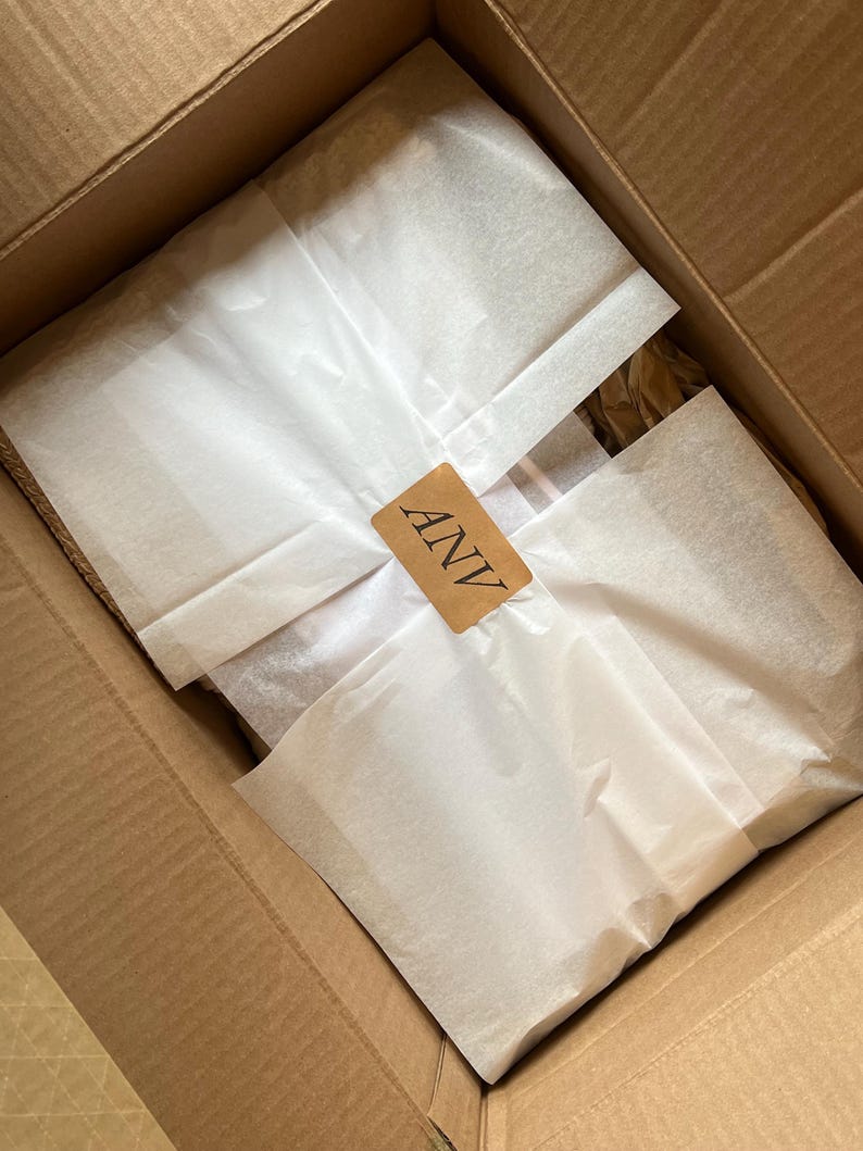 K&ouml;nnte beinhalten: Ein offener Karton mit verpackten Artikeln. Die Artikel sind in wei&szlig;es Seidenpapier eingewickelt und mit einem kleinen braunen Etikett mit der Aufschrift "ANV" versehen.