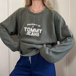 May include: An olive green sweatshirt with the text "PROPERTY OF TOMMY JEANS" in white. The sweatshirt is paired with navy blue athletic shorts with white stripes and the Adidas logo.