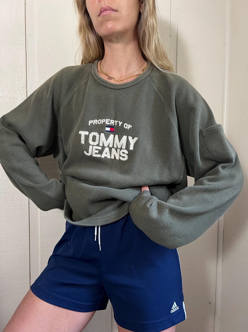 May include: An olive green sweatshirt with the text "PROPERTY OF TOMMY JEANS" in white. The sweatshirt is paired with navy blue athletic shorts with white stripes and the Adidas logo.