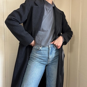 May include: A dark gray wool coat with a classic lapel collar. The coat is worn over a gray turtleneck and light blue denim jeans. The coat is a long length, reaching the ankles.
