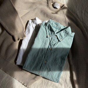 K&ouml;nnte beinhalten: Ein beigefarbener Rundhals-Pullover, ein wei&szlig;es Hemd und ein gr&uuml;n-wei&szlig; gestreiftes Button-Down-Hemd liegen auf einer strukturierten Oberfl&auml;che. Die Kleidungsst&uuml;cke sind gefaltet und erzeugen eine l&auml;ssige, entspannte &Auml;sthetik.