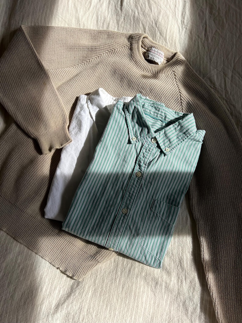 K&ouml;nnte beinhalten: Ein beigefarbener Rundhals-Pullover, ein wei&szlig;es Hemd und ein gr&uuml;n-wei&szlig; gestreiftes Button-Down-Hemd liegen auf einer strukturierten Oberfl&auml;che. Die Kleidungsst&uuml;cke sind gefaltet und erzeugen eine l&auml;ssige, entspannte &Auml;sthetik.