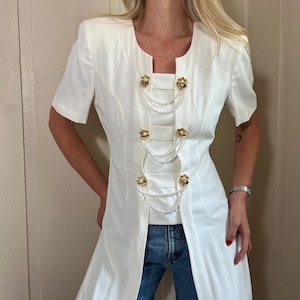 May include: A white short-sleeved dress featuring a square neckline and a front opening. The garment is adorned with three pairs of gold-toned flower-shaped buttons connected by draped white faux pearl chains across the center panel.
