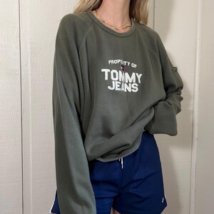 May include: An olive green long-sleeve sweatshirt with the text "PROPERTY OF TOMMY JEANS" in white. Paired with navy blue athletic shorts with white stripes. The outfit is casual and sporty.