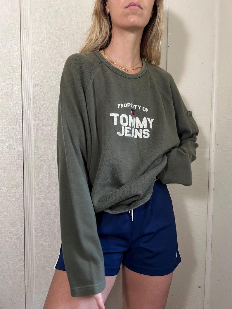 May include: An olive green long-sleeve sweatshirt with the text "PROPERTY OF TOMMY JEANS" in white. Paired with navy blue athletic shorts with white stripes. The outfit is casual and sporty.