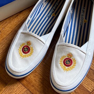 May include: A pair of white canvas slip-on shoes with blue trim and a gold emblem. The interior has blue and white striped fabric. The shoes are new and unworn.