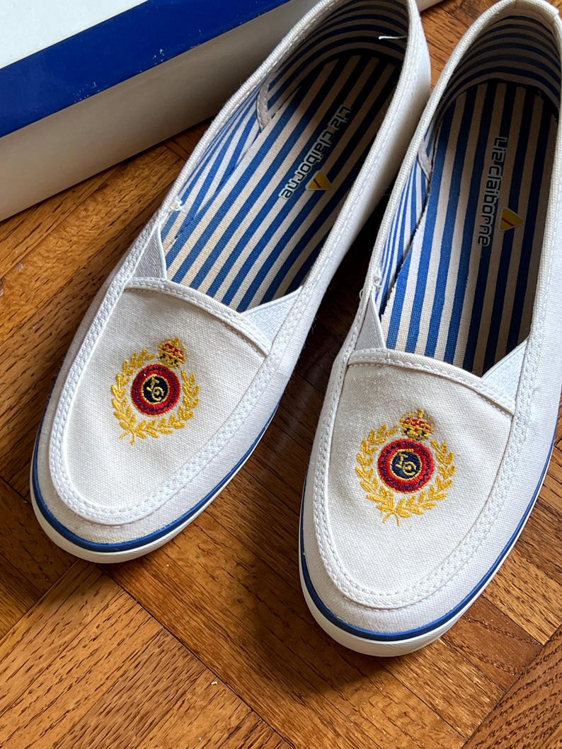 May include: A pair of white canvas slip-on shoes with blue trim and a gold emblem. The interior has blue and white striped fabric. The shoes are new and unworn.