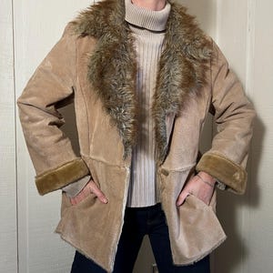 May include: A tan suede jacket with a faux fur collar and cuffs. The jacket has a button closure and two front pockets. The model is wearing a beige turtleneck and dark blue jeans. The jacket is a neutral color and appears to be vintage.