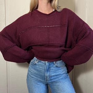 Puede incluir: Un suéter de punto burdeos con cuello en V y un diseño calado horizontal en el pecho. El suéter tiene mangas largas y se combina con unos vaqueros de mezclilla azul claro.