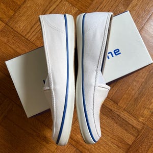 May include: A pair of white canvas slip-on shoes with blue trim. The shoes are new and appear unworn. The soles are white, and the shoes are placed next to a white box with blue text.