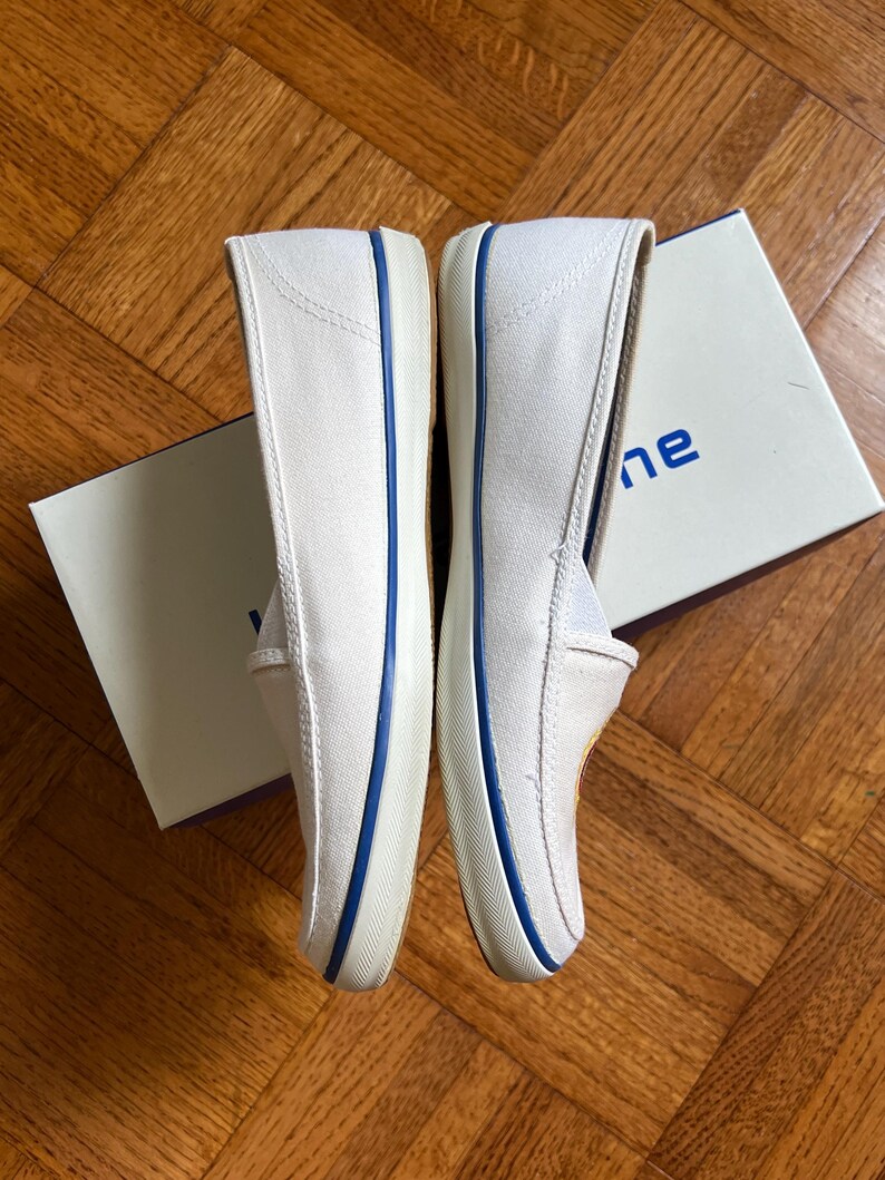 May include: A pair of white canvas slip-on shoes with blue trim. The shoes are new and appear unworn. The soles are white, and the shoes are placed next to a white box with blue text.