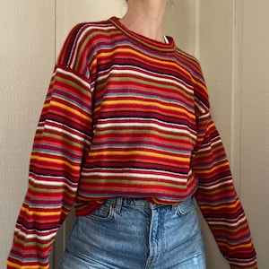 May include: A crew neck sweater with horizontal stripes in red, yellow, green, blue, and white. The long-sleeved sweater is paired with light blue denim jeans. The sweater's stripes create a vibrant, colorful pattern.