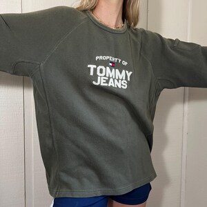 May include: Olive green sweatshirt with the text "PROPERTY OF TOMMY JEANS" in white. The sweatshirt has a crew neck and a small pocket on the upper arm. The Tommy Hilfiger logo is above the text.