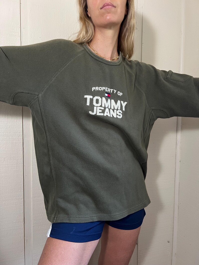 May include: Olive green sweatshirt with the text "PROPERTY OF TOMMY JEANS" in white. The sweatshirt has a crew neck and a small pocket on the upper arm. The Tommy Hilfiger logo is above the text.