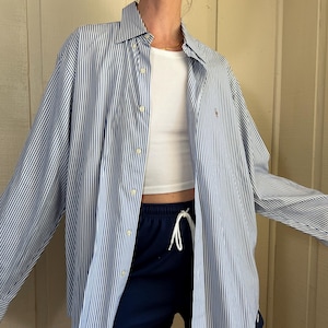 May include: A blue and white striped button-down shirt with long sleeves. The shirt is worn over a white ribbed tank top and navy blue shorts. The shirt has a small logo on the chest.