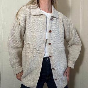 May include: Cream-colored, button-down cardigan sweater with a textured knit pattern. The sweater features a collared neckline, long sleeves, and brown buttons. A unique design element includes a cluster of small, raised knit balls on the front.