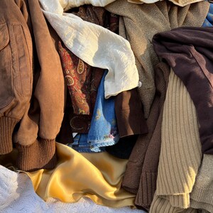 K&ouml;nnte beinhalten: Ein Haufen verschiedener Kleidungsst&uuml;cke in verschiedenen Texturen und Farben. Enth&auml;lt eine braune Wildleder-Bomberjacke, eine cremefarbene Bluse, einen goldenen Satinstoff und einen braunen Strickpullover. Weitere Artikel sind Jeans und gemusterte Stoffe.