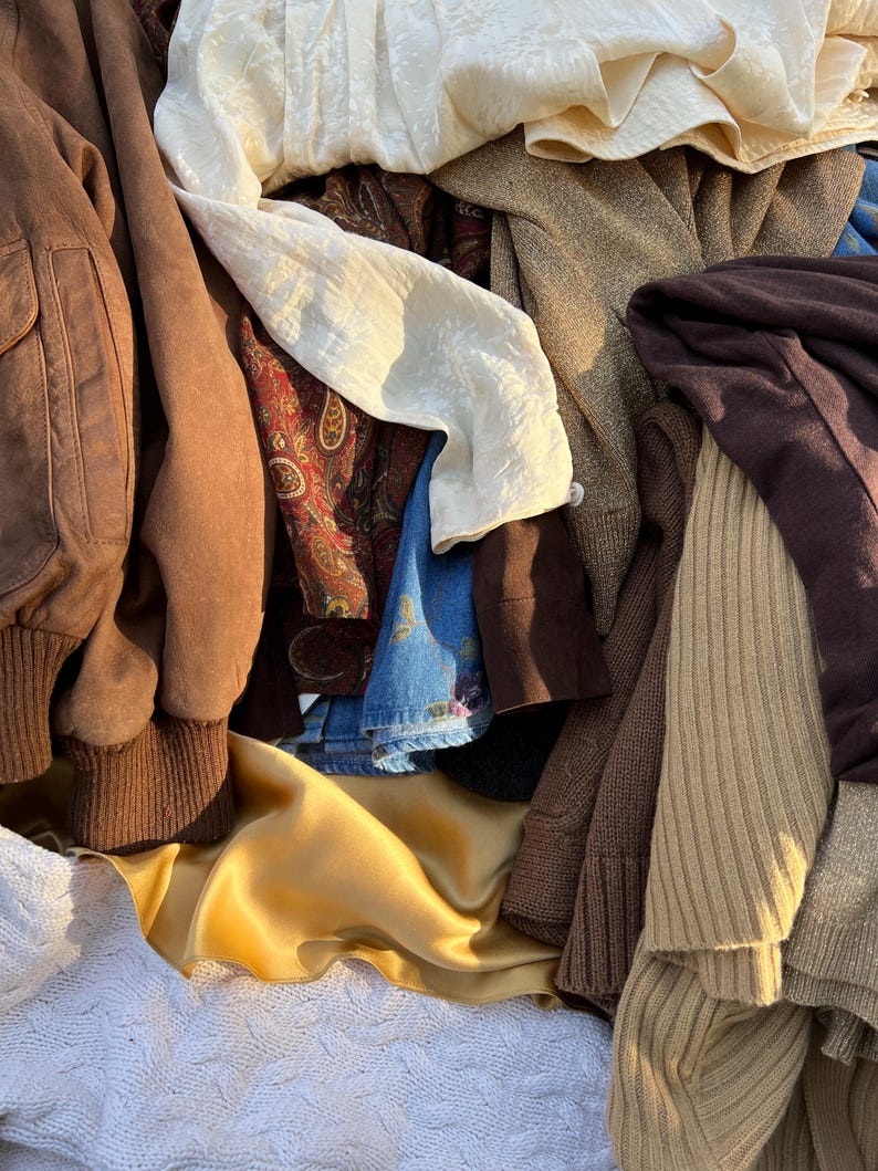K&ouml;nnte beinhalten: Ein Haufen verschiedener Kleidungsst&uuml;cke in verschiedenen Texturen und Farben. Enth&auml;lt eine braune Wildleder-Bomberjacke, eine cremefarbene Bluse, einen goldenen Satinstoff und einen braunen Strickpullover. Weitere Artikel sind Jeans und gemusterte Stoffe.