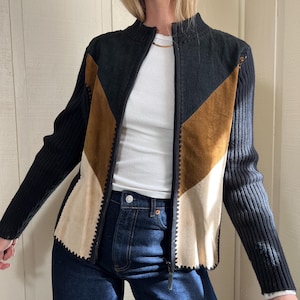 以下が含まれることがあります： 黒、茶色、クリーム色のジップアップジャケット。 ジャケットは、リブ編みの襟と袖を備えた幾何学模様のデザインです。 フロントパネルはスエードとニット素材の組み合わせです。 モデルは白いトップスとブルージーンズを着用しています。