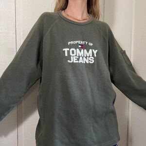 May include: Olive green sweatshirt with the text "PROPERTY OF TOMMY JEANS" in white block letters. The sweatshirt has long sleeves and a crew neck. A small flag graphic is above the text. The model is wearing navy blue shorts.